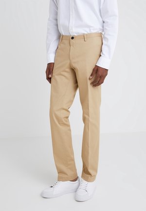 Mand iført beige bukser, hvid langærmet skjorte og hvide sneakers, stående mod en simpel lys baggrund.