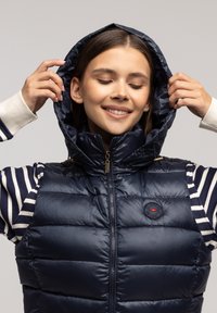 Granatowa kamizelka puchowa z zamkiem z przodu i kapturem. Posiada poziome przeszycia oraz naszywkę z logo na klatce piersiowej. Pod spodem włożony jest paskowany golf.