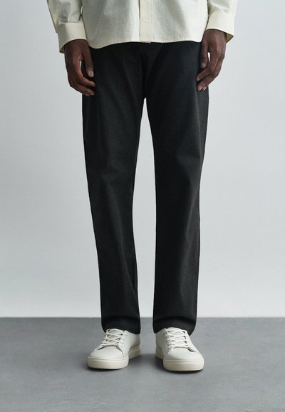 Pantalons noirs effilés avec une texture subtile, associés à des baskets lisses de couleur claire. La chemise est de couleur crème avec un design à boutons.
