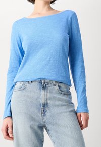 Jasnoniebieska bluzka z długim rękawem o dekolcie w łódkę, uszyta z teksturowanej tkaniny. Połączona z jasnoniebieskimi dżinsami o wysokim stanie.