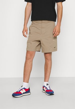 Shorts couleur sable avec une poche latérale zippée et un petit logo. Portés avec des chaussures de sport bleu, rouge et blanc, et un t-shirt noir.