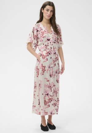 Femme debout portant une robe longue boutonnée à fleurs rose clair avec manches courtes et chaussures noires sans lacets, mains dans les poches.
