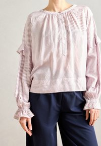 Femme portant un chemisier à manches longues rayé rose clair avec des détails à volants et un pantalon large bleu marine sur un fond uni.