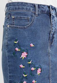 Denimkjol i blått med blommönsterbroderi i rosa och grönt, fem fickor och midjig passform, med klassiska sömnadsdetaljer.