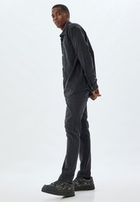 Camisa de mezclilla negra con frente abotonado, combinada con jeans oscuros y zapatillas negras texturizadas. El modelo está de pie de lado contra un fondo claro.