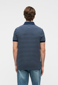 Πόλο μπλούζα σε ναυτικό μπλε χρώμα με λευκές οριζόντιες ρίγες, κοντά μανίκια και γιακά. Το ύφασμα φαίνεται λείο, με ευθεία κοπή στο τελείωμα.