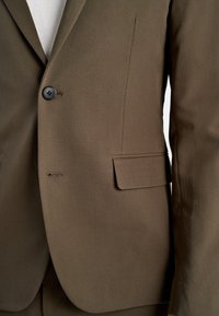 Brauner, maßgeschneiderter Anzugjackett mit einem einzigen schwarzen Knopf, der über einem weißen Hemd befestigt ist, und zeigt eine Klapptasche auf der rechten Seite.