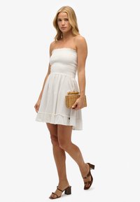 Biała sukienka bez ramiączek z marszczonym topem, rozkloszowaną spódnicą i falbaną na dole. Modelka trzyma plecioną torebkę i ma na sobie brązowe sandały na słupku.