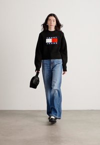 Svart strikkegenser med en dristig farget logo, lys blå vide jeans og svart håndveske; modellen har på seg svart og hvitt fottøy.