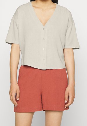 Femme portant un chemisier beige clair à manches courtes avec boutons et col en V et un short taille haute couleur rouille, debout les bras détendus.