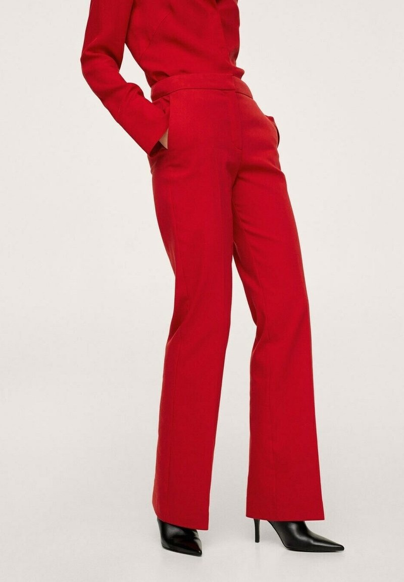 Femme portant un pantalon rouge taille haute et un blazer assorti, les mains dans les poches, associée à des bottines noires à bout pointu et à talons.