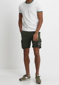 Valkoinen lyhythihainen t-paita, jossa on vihreät reunat, yhdistettynä oliivinvihreisiin cargo-shortsseihin, joissa on taskuja ja paikka. Asusteena harmaat sneakersit.