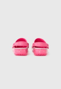Ružičaste Crocs klompe s sjajnom završnom obradom, sa trakicom za sigurnije prianjanje i uzdignutim točkicima na potplatu za bolje prianjanje. Akcenti uključuju zeleni rub.