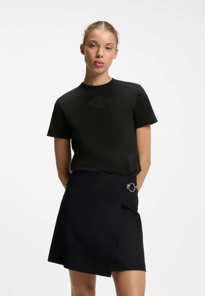 Junge Frau mit lockigem Haar, die ein schwarzes Kurzarmshirt und einen schwarzen Wickelrock mit silbernem Ringdetail trägt, steht vor weißem Hintergrund.
