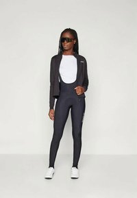 Mustat pyöräilytakki, valkoinen paita ja mustat legginsit kiiltävällä pinnalla. Mustat aurinkolasit ja valkoiset lenkkarit. Yksinkertainen, vartalonmyötäinen muotoilu.