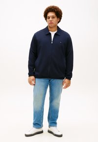 Σκούρο μπλε ζακέτα με φερμουάρ, γιακά που στέκεται όρθιος, μακριά μανίκια και πλαϊνές τσέπες. Συνδυάζεται με ανοιχτό μπλε τζιν και λευκά αθλητικά παπούτσια.