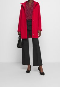 Rode hooded jas met een ritssluiting, gebloemd blouse, zwarte wijde broek en zwarte stilettos. Houdt een zwarte handtas vast.