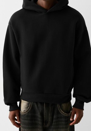 Person iført en sort hættetrøje med en forlomme og løstsiddende ærmer, kombineret med mørke, falmede jeans med synlige sømme.