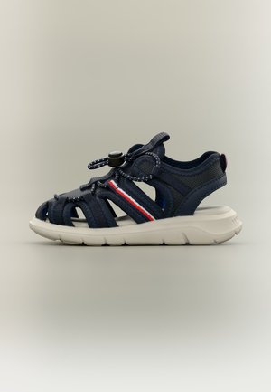 Marineblå sportsandal med hvit såle, justerbar stropp med spenne, mesh-sider, og rød-hvit-blå stripet detalj på siden.