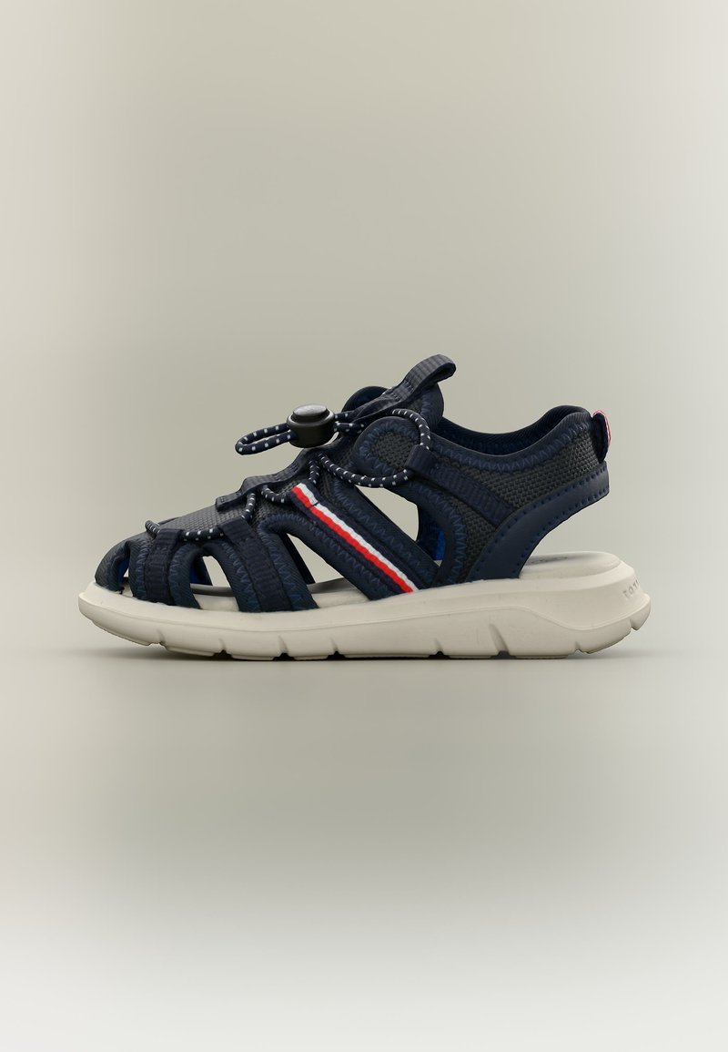 Αθλητικό σανδάλι σε ναυτικό μπλε χρώμα με λευκή σόλα, ρυθμιζόμενο λουράκι με κουμπί, πλευρές από δίχτυ και λεπτομέρειες με κόκκινες, λευκές και μπλε ρίγες στο πλάι.