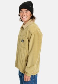 Giacca in corduroy di un beige chiaro con colletto in pelliccia sintetica, dotata di tasca sul petto e toppa con logo, indossata con jeans blu e un berretto nero.