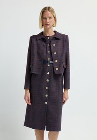 Mujer vistiendo un vestido midi abotonado en azul oscuro y burdeos con una chaqueta corta a juego y botones dorados, de pie frente a un fondo liso.