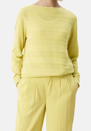 Personne portant un pull tricoté jaune clair avec des motifs ajourés horizontaux et un pantalon ample assorti, les mains dans les poches.