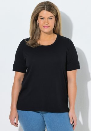 Frau mit schulterlangem hellbraunem Haar, trägt Creolen, ein schwarzes kurzärmeliges Shirt und blaue Jeans, steht vor einem weißen Hintergrund.