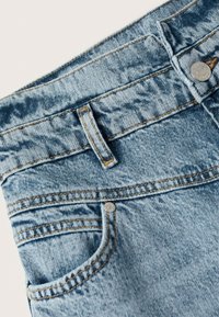 Jeans de denim azul claro con costuras doradas, que cuentan con un bolsillo frontal, un cierre de botón y una superficie ligeramente texturada.