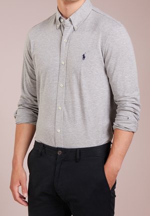 Mand iført lysegrå skjorte med knapper og opkrogede ærmer samt sorte bukser, stående med den ene hånd i lommen mod en ensfarvet baggrund.