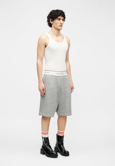 Λεπτός άνδρας με σγουρά μαλλιά φοράει λευκό αμάνικο φανελάκι, γκρι μακριά σορτς, ριγέ κάλτσες και μαύρα μποτάκια από πατέντα δέρμα, στέκεται σε ουδέτερη στάση.