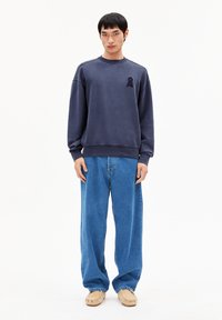 Sweat-shirt bleu marine avec un col rond, des poignets et un ourlet côtelés ; présente un emblème sombre texturé. Associé à un jean bleu clair ample.