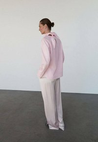 Chemisier rose pâle à manches longues avec un nœud au dos, associé à un pantalon large crème, sur un fond neutre. Textures lisses visibles.