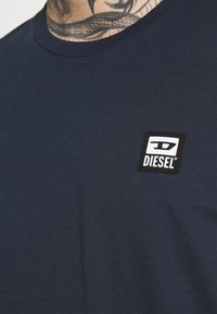 Närbild av en mörkblå Diesel-t-shirt med en fyrkantig logotyp-patch på bröstet och delvis vy av en nacktattoo.