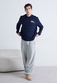 Tamsiai mėlynas ilgomis rankovėmis marškinėliai su Lacoste logo ir pilki sportiniai kelnių su šoninėmis kišenėmis bei elastiniais rankogaliais, padėti ant šviesiai pilko kilimo.