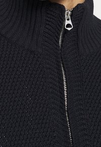 Pull noir en maille avec col montant et fermeture éclair argentée. Présente un motif de point texturé et des détails en tricot côtelé au niveau du col.