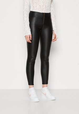 Personne portant un legging en cuir noir taille haute avec une fermeture éclair à l'avant, un top blanc en dentelle à manches longues, et des baskets blanches avec un logo swoosh pêche.