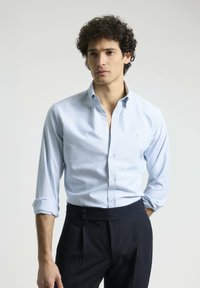 Camisa de rayas azul claro con cuello abotonado, puños enrollados y botones blancos, combinada con unos pantalones oscuros a medida con detalles de botones.