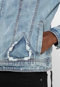 Hellblaue Jeansjacke mit einem dreieckigen, ausgefransten Fleck nahe der Taille und einer Hand in der rechten Seitentasche, getragen über einer schwarzen Hose.