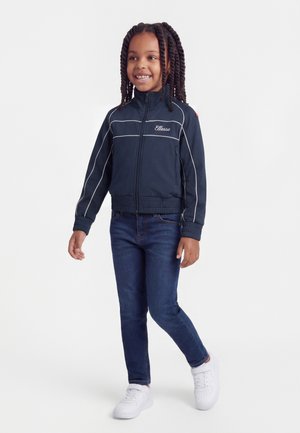 Ragazza sorridente con capelli intrecciati che indossa una giacca blu navy con cerniera e bordi bianchi, jeans scuri e scarpe da ginnastica bianche, in piedi davanti a uno sfondo bianco.