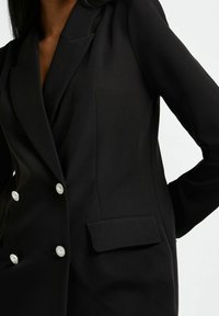 Černý blazer s dvouřadým zapínáním, vyrobený z hladké tkaniny. Obsahuje kulaté knoflíky ve tvaru perel a jednu kapsu s klopou na přední straně.