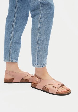 Sandalias de cuero rosa con diseño de tiras cruzadas y detalles en hardware dorado. Suela de corcho que aporta textura. Combinadas con pantalones vaqueros azul claro.