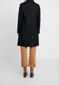 Osoba v černém kabátě, zastrčených hnědých kalhotách a kotníkových botách na vysokém podpatku se vzorem hadí kůže, stojící před jednobarevným pozadím.