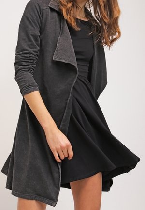 Veste longue à fermeture ouverte en tissu gris foncé, avec une finition texturée et des manches roulées, portée sur une robe noire à ourlet fluide.
