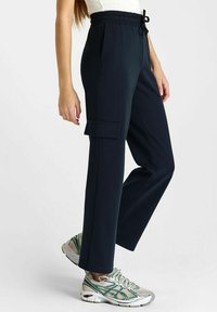 Vrouw in marineblauwe cargobroek met elastische tailleband en witte sneakers, staand tegen een effen achtergrond.