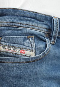 Jeans de denim azul com uma textura desgastada, apresentando um rótulo proeminente no bolso de trás a dizer "Diesel Industry Denim Division" e detalhes visíveis de costura.