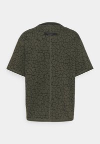 Camisa de manga corta en verde oliva con un patrón agrietado. Presenta un cuello redondo y un corte holgado, hecha de una tela texturizada.