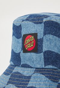 Chapéu bucket xadrez azul feito de denim, com um patch do logotipo "Santa Cruz" em vermelho e preto. Aba costurada com detalhes amarelos.