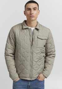 Quilted beige jakke med glidelås foran, krage og en brystlomme. Har avrundede søm mønstre og en glatt tekstur.