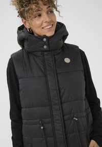 Čierna puffer vesta s vysokým golierom, cvočkovými gombíkmi a detailmi s elastickým šnurovaním. Má prešívanú textúru a logo značky na hrudi.